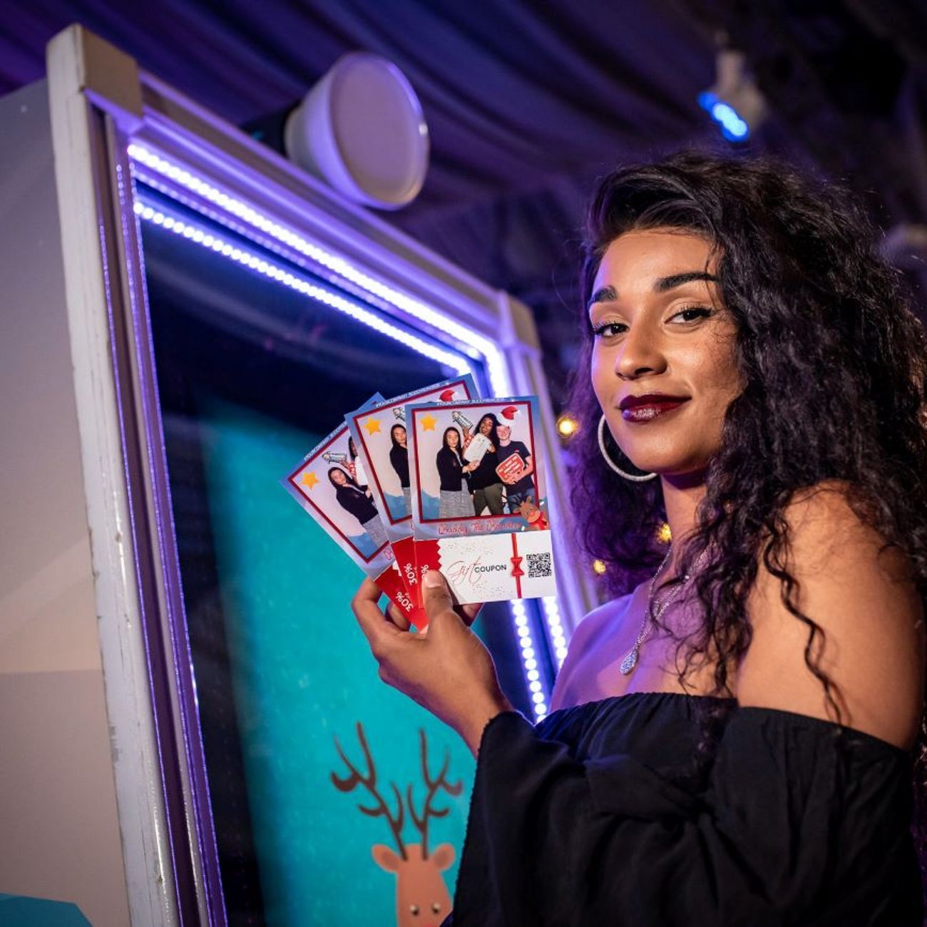 Guest holding up four custom-branded photo prints next to the magic mirror at a Christmas event, LED frame illuminated and dye-sublimation printer visible on the booth