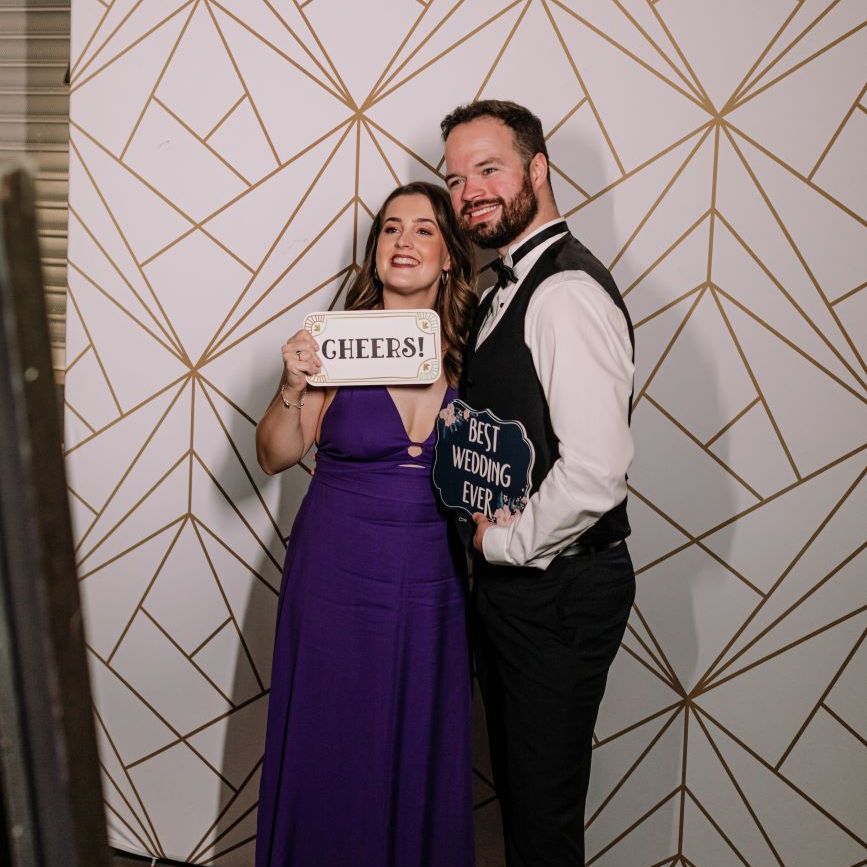 Wedding guests posing at the magic mirror with 'Cheers!' and 'Best Wedding Ever' prop signs, geometric gold-on-white backdrop behind them