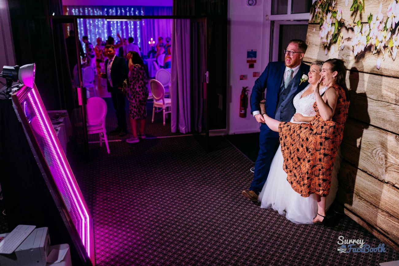 Bride and groom posing at the magic mirror with the pink-LED frame fully illuminated, captured photo preview visible on the mirror's screen