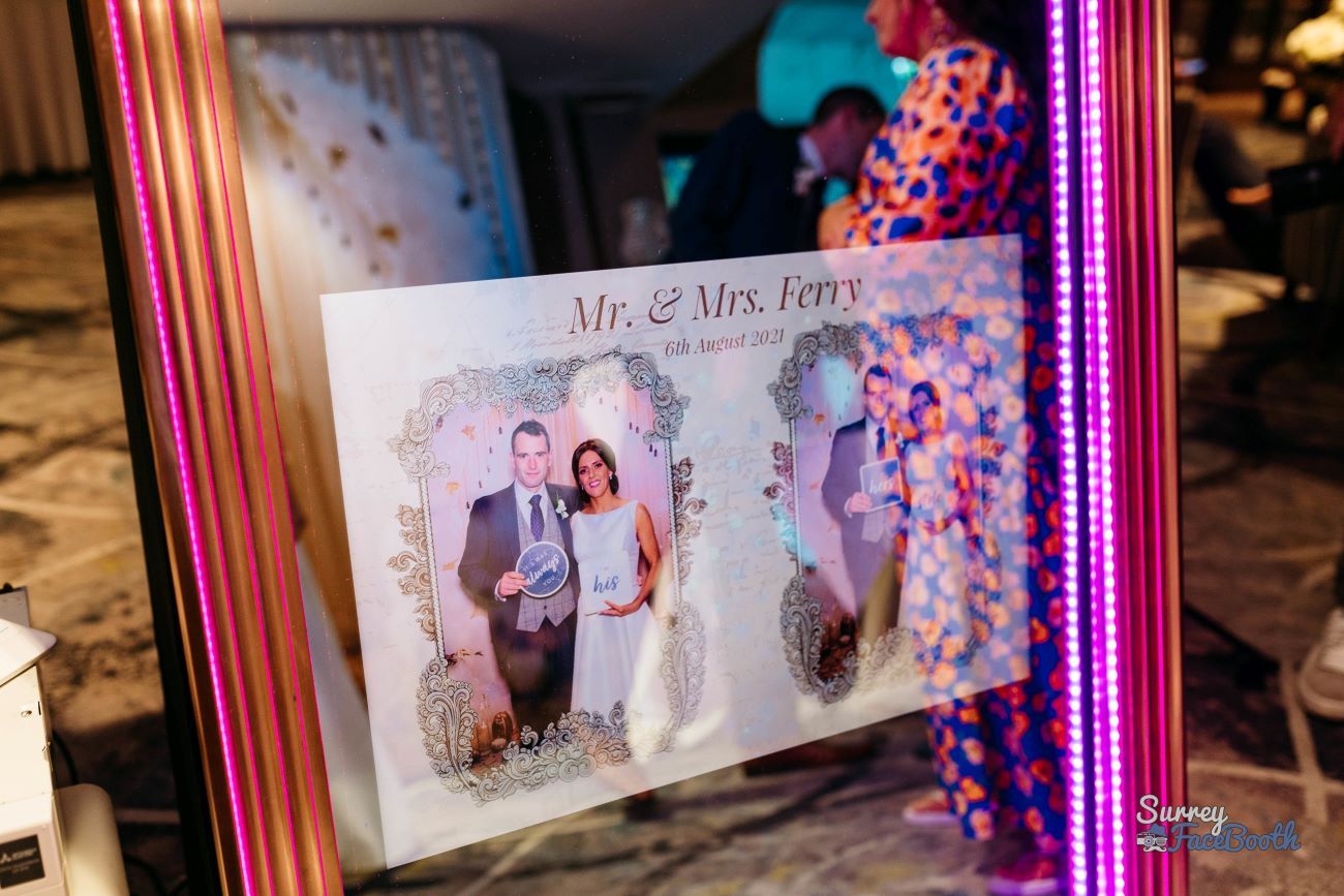 Wedding photo print with a custom 'Mr. & Mrs. Ferry, 6th August 2021' overlay design, held up in front of the magic mirror's pink LED frame