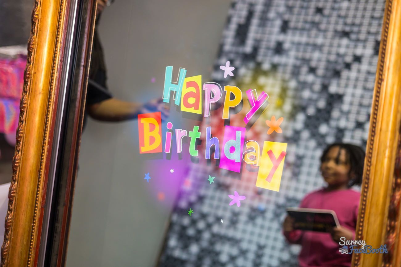 Close-up of the magic mirror's touchscreen showing a colourful animated 'Happy Birthday' overlay, gold mirror frame around the edge with a child reflected at the side