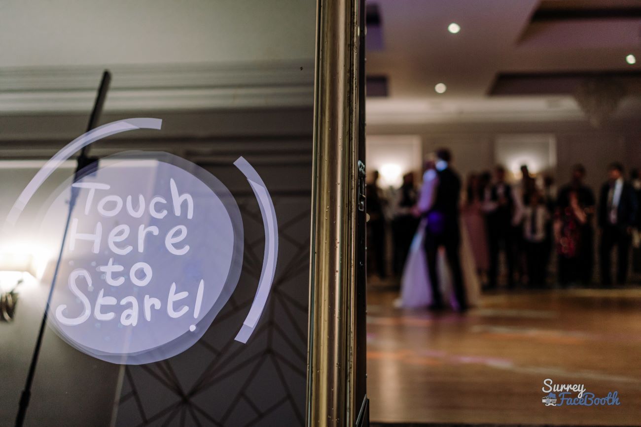 Close-up of the magic mirror's 'Touch Here to Start' animated prompt, with the bride, groom and wedding guests gathered on the dance floor reflected in the mirror behind