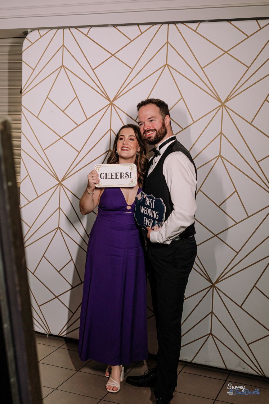 Wedding guests posing at the magic mirror with 'Cheers!' and 'Best Wedding Ever' prop signs, geometric gold-on-white backdrop behind them